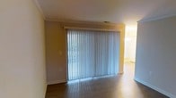 This single-story space presents a clean, functional layout with notable architectural finishing touches. The living area is a standout feature, highlighted by a large sliding glass door dressed with full-length vertical blinds that filter natural light into the room, creating a bright and airy atmosphere. The wide door frame is accented with crown molding trim, adding a polished, traditional character to the space.

The flooring throughout is a rich, dark hardwood-style material that contrasts neatly with the neutral beige walls and crisp white trim, including baseboards and crown molding that run consistently through the visible areas. The ceiling carries additional crown molding detailing, reinforcing a sense of refined craftsmanship.

An adjacent area, visible through an open passageway, suggests a connected dining or kitchen space, illuminated by a decorative light fixture that adds warmth to the overall interior. The open sightlines between rooms contribute to a cohesive, flowing layout. Overall, the space offers a well-maintained interior with classic design elements and practical features suited for comfortable everyday living.