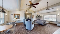 Located at 3240 Germantown Rd in Memphis, TN, this single-floor clubhouse-style space offers a thoughtfully designed interior defined by soaring vaulted ceilings with exposed beam detailing and an open, airy atmosphere throughout. The main gathering area is anchored by a stone fireplace flanked by large windows that admit generous natural light and offer views of the surrounding landscape. Ceiling fans and a combination of pendant chandeliers and recessed lighting provide both comfort and ambiance across the space.

The lounge area is organized into distinct seating zones, featuring plush sofas, upholstered accent chairs, and coffee tables arranged atop a richly patterned carpet that contrasts with hardwood-style flooring in transitional areas. A dedicated dining nook accommodates a round table with cross-back chairs, accompanied by pendant lighting and a decorative mirror. Framed artwork and wall sconces contribute to the refined yet welcoming aesthetic.

The kitchen features dark granite countertops, a stainless steel sink, light cabinetry, and a full-size refrigerator, with a pass-through window connecting it to the main area. Additional rooms include a hallway, bathroom, closet, and a flexible-use space, completing this well-rounded and versatile communal environment.