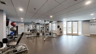 Located at 91 York Rd in Willow Grove, PA, this fitness center offers a generously sized, single-floor workout facility designed to support a well-rounded exercise routine. The space features durable speckled rubber flooring throughout, providing both safety and comfort underfoot across all workout zones.

The strength training area is well-equipped with a multi-functional cable machine, adjustable benches, a full dumbbell rack, and a selection of stability balls and kettlebells neatly organized on dedicated racks. Wall-mounted mirrors in this zone enhance the sense of space and allow for proper form monitoring during workouts.

The cardio section includes upright bikes, treadmills, and an elliptical trainer, arranged beneath motivational wall lettering that reads "FITNESS SWEAT HYDRATE." Wall-mounted flat-screen televisions are positioned throughout for entertainment during workouts.

Natural light filters in through multiple windows and glass double doors that open to an outdoor view, creating a bright and inviting atmosphere. Recessed lighting in the drop-panel ceiling ensures consistent illumination throughout. Additional amenities include a water fountain and a hand sanitizer station, supporting hygiene and hydration. Motivational fitness artwork adorns the light gray walls, contributing to an energizing and focused environment suitable for a wide range of fitness activities.