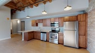 This loft-style unit masterfully combines the raw character of an industrial conversion with thoughtfully updated modern finishes. The open living area makes a striking impression with soaring ceilings showcasing exposed heavy timber beams and visible ductwork, evoking the building's historic origins. Original brick masonry walls in warm tan tones run throughout the space, punctuated by large, black-framed arched and grid windows that flood the interior with abundant natural light and offer views of the surrounding urban streetscape.

Light hardwood floors flow seamlessly through the main living area and into the kitchen, which is equipped with warm-toned wood cabinetry, light countertops, a peninsula breakfast bar, and a full suite of stainless steel appliances including a refrigerator, range, over-the-range microwave, and dishwasher. Industrial-style pendant lighting reinforces the loft aesthetic throughout. The bedroom continues the brick wall detailing, complemented by soft carpeting and generous natural light from the large windows. The bathroom offers a clean, contemporary finish with tile flooring, a tub-shower combination, and a wood-accented vanity with a light countertop. The overall ambiance is one of urban sophistication, balancing historic architectural texture with fresh, functional living spaces.