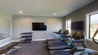 This fitness center is designed to support a comprehensive workout experience within a single, open floor plan. The space is finished with neutral gray walls, recessed lighting, and a combination of carpet and rubber flooring that defines distinct activity zones. Multiple wall-mounted flat-screen televisions are positioned throughout, providing entertainment during cardio sessions.

The cardio zone features a row of Matrix treadmills and an elliptical trainer, positioned in front of windows that allow natural light and outdoor views. Adjacent to the cardio area, a dedicated free weights section includes tiered dumbbell racks stocked with a full range of weights, a flat workout bench, and a large wall mirror that enhances the sense of space.

A functional training zone houses a multi-station cable and strength machine, stability balls in various sizes, medicine balls, and resistance bands organized on a standing rack. A full-length glass partition wall separates the fitness center from an adjoining lounge or amenity space, offering visual connectivity while maintaining acoustic separation. The overall design balances practicality with a clean, modern aesthetic suited for a variety of fitness routines.