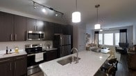 This single-floor residence offers a thoughtfully designed open-concept layout that seamlessly connects the kitchen and living areas. The kitchen is a standout feature, showcasing rich dark cabinetry that provides a bold contrast against the light granite countertops. A spacious kitchen island with an undermount stainless steel sink and elegant pendant lighting serves as the centerpiece of the space. Stainless steel appliances, including a French door refrigerator, electric range, and built-in microwave, complete the contemporary kitchen setup. Track lighting overhead ensures the cooking area is well-lit and functional. The open flow from the kitchen leads directly into a comfortable living area, which benefits from natural light streaming through large windows dressed with blinds. A ceiling fan adds to the comfort of the living space. The overall ambiance of the residence is modern and sophisticated, with dark wood-toned flooring and a cohesive color palette of deep espresso, cool grays, and crisp whites that lend the space a clean, upscale aesthetic. The thoughtful integration of lighting, materials, and layout makes this a well-appointed and highly livable space.