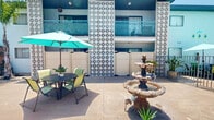 Located at 2635 Milton Ave in Fullerton, California, this mid-century modern residential complex showcases a distinctive architectural style defined by bold teal and white exterior tones, decorative breeze-block screen panels, and diamond-patterned balcony railings that lend the property a retro yet refreshed aesthetic.

At the heart of the complex lies a spacious open-air courtyard, thoughtfully designed for community living. Tall palm trees provide natural shade and a Southern California ambiance, while a classic multi-tiered stone fountain serves as a charming focal point. The courtyard features a dedicated lounge area complete with wicker furniture and a built-in outdoor fireplace, as well as umbrella-shaded dining tables ideal for outdoor gatherings. A swimming pool is also accessible from the courtyard, adding to the resort-like atmosphere.

A covered gazebo area functions as a shared communal space, furnished with a large wicker dining set and adorned with decorative wall art. Rows of individual resident mailboxes line the walls beneath the gazebo, combining convenience with community. Ground-floor units open directly onto the courtyard through sliding glass doors and private entrances, while upper-level units enjoy balcony access. The complex benefits from abundant natural sunlight throughout, characteristic of its Fullerton, CA location.