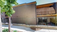 This single-story property presents a well-maintained exterior characterized by a smooth stucco finish in a warm earth tone, complemented by a dark trim along the roofline that adds a clean, modern contrast. A sturdy metal railing fence borders the front of the building, offering both a defined boundary and an open, airy feel to the entrance area. The ground-level entry features a gated access point, providing a sense of security while maintaining visual openness.

One of the standout features of this property is the landscaped side walkway, which is lined with mature, leafy trees that provide generous shade and a canopy of greenery overhead. Beneath the trees, a variety of flowering plants and ground-level shrubs add vibrant color and texture to the pathway, creating an inviting and pleasant outdoor corridor. The walkway is paved with clean concrete, ensuring easy and accessible passage along the side of the building.

An upper-level balcony is visible on an adjacent structure, fitted with a natural bamboo or reed screen that adds a warm, organic element to the overall aesthetic. The outdoor areas appear bright and sun-drenched, consistent with a warm, sunny climate. Together, the architectural details and thoughtfully maintained landscaping create a welcoming and functional outdoor environment that enhances the overall character of the property.