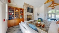 Located at 36916 Crooked Hammock Way in Lewes, Delaware, this single-floor community space offers an open-concept layout that seamlessly integrates a living room, dining area, and kitchen into one cohesive and inviting environment. The living room is anchored by a dramatic floor-to-ceiling stacked stone fireplace, topped with a wall-mounted television and flanked by warm wood built-in bookshelves with glass-front cabinetry. Soaring vaulted ceilings with exposed wood beams lend an airy, coastal character throughout, while large arched and multi-pane windows flood the interior with natural light and offer direct views of an outdoor pool. The dining area features multiple round marble-top tables paired with woven cane-back chairs, all set beneath the same high vaulted ceiling. Patterned tile-style carpeting flows throughout the main gathering areas, complementing the neutral, coastal-inspired palette. The kitchen is outfitted with rich warm-toned wood cabinetry, a mosaic tile backsplash, granite countertops, a built-in wine rack, and pendant lighting over a spacious bar-height counter with seating. An arched hallway leads to additional rooms, completing a well-designed and functional space ideal for community gatherings.