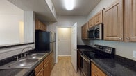 This single-floor residence offers a thoughtfully designed layout that balances functionality and comfort throughout. The galley-style kitchen is a standout feature, outfitted with warm honey-toned cabinetry, dark granite countertops, and a full suite of stainless steel appliances including a smooth-top electric range, over-the-range microwave, and a refrigerator. A double stainless steel sink adds practicality, while light wood-look flooring and a textured ceiling complete the space. The kitchen connects seamlessly to the rest of the home through a doorway leading to the interior hallway. The living area is generously proportioned, featuring plush neutral carpeting, a ceiling fan with light fixture, and a window that allows natural light to filter in. An adjoining dining or flex space transitions between tile and carpet flooring, with a pendant light overhead and additional window views. The bathroom features rich dark-toned cabinetry, granite countertops, a large vanity mirror illuminated by a multi-bulb light bar, and a window providing natural light. A dedicated in-unit laundry area provides added convenience with a full-size washer and overhead storage shelf. The living space also benefits from a door with adjacent windows that opens to the outdoors, enhancing the sense of openness and connectivity to the exterior.