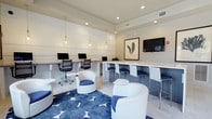 This contemporary single-floor space is thoughtfully designed to serve as a functional yet stylish business center or collaborative lounge. The room features a clean, modern aesthetic anchored by a striking blue-and-white color scheme that runs throughout the furnishings and décor.

Along one wall, a sleek white workstation counter houses multiple desktop computers with ergonomic mesh task chairs, providing a dedicated area for focused work. Pendant Edison bulb lights hang above, casting warm, ambient light that complements the recessed ceiling lighting throughout the space.

Adjacent to the workstations, a continuous high-top white counter runs along the wall, paired with modern white-and-blue counter-height chairs. This bar-style seating area is ideal for casual meetings or independent work. A flat-screen television is mounted on the wall nearby, alongside a framed botanical art piece that adds a subtle natural touch.

The lounge area features curved white accent chairs with blue cushions arranged around small round side tables, encouraging informal gatherings or relaxed breaks. A bold blue area rug with a geometric star pattern anchors this seating area, adding visual interest to the light-toned tile flooring. The white paneled walls and tray ceiling details lend an air of refined sophistication to the overall environment.