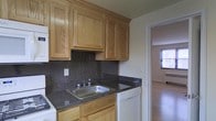 This single-floor space features a well-appointed kitchen as a central highlight. The kitchen is outfitted with warm oak cabinetry, providing generous upper and lower storage. Dark speckled granite countertops complement the wood tones and extend across the workspace, while a matching granite backsplash adds a cohesive, polished look. A stainless steel sink is positioned beneath the cabinetry, and a white dishwasher is integrated seamlessly into the layout. A range is also present, completing the functional cooking setup. The kitchen opens through a doorway into an adjoining room, which features hardwood flooring that lends warmth to the space. Natural light filters in through a window visible from the kitchen, brightening the connected area and contributing to an overall airy feel. The neutral wall tones throughout complement both the kitchen finishes and the hardwood floors, creating a clean and practical living environment. The single-floor layout ensures convenient and accessible flow between areas.