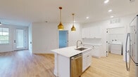 Located at 7015 Ruth Ave in Wilmington, NC, this well-appointed single-story home offers three bedrooms, two bathrooms, and a thoughtfully designed layout. The heart of the home is an open-concept kitchen and living area finished with light hardwood flooring throughout. The kitchen is a standout feature, boasting crisp white shaker cabinetry with brushed gold hardware, a white quartz center island with a stainless steel sink, subway tile backsplash, and a full suite of stainless steel appliances including a refrigerator, range, microwave, and dishwasher. Striking gold pendant lights hang above the island, adding a modern, elegant touch. The living area flows seamlessly from the kitchen, offering a bright, open atmosphere. The spacious primary bedroom provides generous room to furnish comfortably, while the additional bedrooms feature plush carpeting, ceiling fans, and natural light from windows with blinds. Hardwood-floored bedrooms offer a clean, contemporary feel. Both bathrooms are finished with white vanities, quartz countertops, and hardwood-style flooring. A dedicated laundry room with a full-size washer and dryer and overhead shelving adds everyday convenience. Clean white walls and consistent finishes create a cohesive, modern aesthetic throughout.