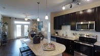 This single-story residence at 811 Spring Forest Rd in Raleigh, NC, offers a thoughtfully designed layout with a seamless flow between living spaces. The kitchen serves as a central highlight, showcasing dark espresso cabinetry, granite countertops, a subway tile backsplash, and stainless steel appliances. A curved kitchen island with pendant lighting overhead provides additional prep space and casual seating, while track lighting adds a modern touch. The kitchen opens toward the living area, which benefits from natural light streaming through large windows and a glass door, creating a bright and welcoming atmosphere.

The floor plan, as seen in the aerial view, reveals two well-proportioned bedrooms, each styled with tasteful furnishings, decorative artwork, and warm, neutral tones. The primary bedroom features coordinated décor with layered bedding and accent pieces that contribute to a refined, comfortable feel. A bathroom is also visible within the layout, appearing to include a soaking tub and vanity area with ample lighting. Throughout the space, warm wood-tone flooring and neutral wall colors create a cohesive, inviting ambiance well-suited to comfortable everyday living.