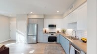 North Grove A1D Model #3 is a thoughtfully designed 620-square-foot, one-bedroom, one-bathroom apartment located in Riverside, CA 92501. The open-concept kitchen and living area serves as the heart of the unit, featuring two-tone shaker cabinetry in soft blue-gray and white, a white subway tile backsplash, light stone countertops, and stainless steel appliances including a French door refrigerator, gas range, built-in microwave, and dishwasher. Light wood-style flooring flows throughout the main living areas, complementing the bright, neutral color palette. The living room receives ample natural light through multiple large windows and offers access to a private covered patio with views of palm trees. The 120-square-foot bedroom is carpeted and includes a large window with outdoor views and direct access to a spacious walk-in closet equipped with wire shelving and dual hanging rods. The bathroom features a blue-gray vanity with an undermount sink, stone countertop, a large mirror, and a full shower-tub combination. A hallway connects all rooms efficiently, completing this well-appointed, single-floor residence ideal for comfortable urban living.