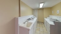This shared laundry facility is a practical, well-organized space designed to accommodate the needs of multiple users. The main laundry area is equipped with a row of numbered washing machines and dryers neatly arranged along the walls, accompanied by utility sinks that provide added convenience for hand-washing or pre-treating laundry. Bright fluorescent lighting illuminates the space, and light tile flooring throughout makes for easy maintenance and a clean appearance.

A separate adjoining room, finished with patterned carpet flooring and a ceiling light fixture, serves as a dedicated folding and sorting area. This room features a long countertop along one wall, ideal for folding garments, as well as a rolling laundry cart for transporting loads with ease. A secure door with a window panel connects this room to other areas of the building.

Completing the amenity space is a Pepsi vending machine, offering refreshments while waiting for laundry cycles to finish. An additional folding counter nearby provides extra workspace. The walls throughout are finished in warm neutral tones, contributing to a functional and welcoming atmosphere. Overall, this facility is thoughtfully equipped to serve as a convenient, communal laundry resource.