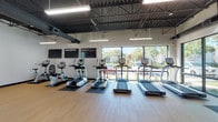 This fitness center is a single-floor facility designed to accommodate a wide range of workout needs within an open, well-organized layout. The space is divided into distinct functional zones, each equipped with purpose-specific machines and equipment. The cardio area features a row of treadmills, ellipticals, and stationary bikes positioned along a wall of large, floor-to-ceiling windows that invite abundant natural light and provide views of the surrounding outdoor greenery. Multiple flat-screen televisions are mounted along the adjacent wall, enhancing the cardio experience.

The strength training area occupies a generous portion of the floor plan, housing a selection of cable machines, weight benches, incline benches, and free weights including dumbbells and medicine balls. Full-length mirrors line one wall, supporting proper form and adding depth to the space. A dedicated section with pink accent walls distinguishes this zone visually while maintaining a cohesive overall aesthetic.

Throughout the facility, durable wood-look flooring provides a clean and resilient surface, complementing the industrial-style exposed ceilings with dark structural beams, visible ductwork, and suspended linear lighting fixtures. The high ceilings contribute to an airy, spacious atmosphere, while large windows across multiple walls ensure consistent natural light and an open, welcoming environment.