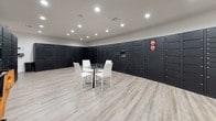 This single-floor package room is purpose-built to streamline parcel management, centered around a comprehensive Luxer One smart locker system. Banks of matte black lockers line the walls on multiple sides, offering a wide range of compartment sizes to accommodate packages of varying dimensions — from small envelopes to larger deliveries. The lockers extend floor to ceiling, maximizing storage capacity while maintaining a sleek, uniform appearance. An integrated digital kiosk is embedded within the locker bank, enabling residents to receive notifications and retrieve their packages securely and efficiently. The room features light wood-look flooring that contrasts cleanly with the dark locker units, contributing to a modern, organized atmosphere. Recessed ceiling lighting provides bright, even illumination throughout the space. A small round glass-top table with white chairs is centrally positioned, offering a convenient surface for sorting or managing deliveries. Bilingual instructional signage is displayed on the locker units, ensuring accessibility for all users. The open floor plan allows for easy navigation between locker banks, and the overall design reflects a deliberate focus on functionality, security, and a polished aesthetic suited to a contemporary residential or mixed-use setting.