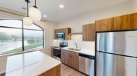 This single-floor unit showcases a contemporary design aesthetic with a cohesive blend of warm and neutral tones throughout. The kitchen serves as a central highlight, featuring flat-front cabinetry in a rich walnut-tone finish paired with crisp white quartz countertops. A full suite of stainless steel appliances includes a refrigerator, electric range, built-in microwave, and dishwasher. A white subway tile backsplash adds texture behind the sink area, while globe pendant lights overhead contribute to a modern, curated look. A kitchen island with matching cabinetry provides additional counter space and storage. Wood-look flooring runs throughout the main living areas, adding warmth and continuity to the space.

A separate room features large, arched-top windows that frame views of the surrounding neighborhood streetscape, allowing generous natural light to fill the space. The bathroom is finished with a walnut-tone vanity, white quartz countertop, a large frameless mirror, and modern vanity lighting, complemented by neutral tile flooring. A panel-style door with classic detailing adds a refined touch. The overall layout, visible in the floor plan, offers a functional and open arrangement with clear separation between the living, kitchen, and private areas.