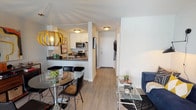This single-floor residence is thoughtfully laid out to maximize functionality and style. The open-concept main living area seamlessly connects the kitchen, dining, and living spaces. A galley-style kitchen features white cabinetry, dark countertops, stainless steel appliances including a microwave and range, and a peninsula with bar seating that doubles as a casual dining spot. Light wood-style flooring runs throughout, lending a cohesive, airy feel to the space.

The living area accommodates a plush navy sectional sofa paired with a glass coffee table, anchored by a bold black-and-white art piece. A decorative orb pendant light adds a striking design element near the dining space. An elegant arched doorway serves as a architectural focal point, connecting the living room to the bedroom.

The bedroom offers a built-in desk nook with open industrial-style shelving, a mirrored closet, and ample room for a seating area, providing a versatile and comfortable retreat. The bathroom includes a full-size tub and shower combination with clean white finishes and chrome fixtures. Throughout, the light-toned walls and consistent flooring create a bright, modern, and cohesive living environment.