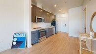 This modern studio in West Sacramento, CA offers a thoughtfully designed open-concept layout that seamlessly integrates the kitchen and living area into a single, cohesive space. Light wood-tone flooring flows throughout, complementing the clean white walls and recessed lighting that give the unit a bright, contemporary feel.

The kitchen is a standout feature, outfitted with sleek gray cabinetry extending to the ceiling for generous storage, light countertops, and a full suite of stainless steel appliances including a refrigerator, range, microwave, and dishwasher. The kitchen flows directly into the open living area, making the space ideal for both everyday living and entertaining.

The living area is bathed in natural light, with a large sliding glass door leading to a private balcony that offers open views of the West Sacramento skyline — a particularly attractive feature of this unit. An additional window further enhances the airy ambiance of the space.

The bathroom is clean and functional, featuring a full soaking tub with shower, and a neutral color palette that maintains the unit's modern aesthetic. A dedicated closet provides practical storage. The overall design is sleek and efficient, making excellent use of the available footprint while maintaining a comfortable, welcoming atmosphere.