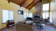 This single-floor space makes a strong architectural impression with its dramatic vaulted ceiling clad in warm wood paneling and supported by exposed wooden beams, creating a sense of generous vertical volume throughout the main living area. The ceiling's steep pitch rises to a commanding height, enhancing the open and airy feel of the space.

The focal point of the room is a full-height brick fireplace, which extends from floor to ceiling and anchors the space with a rustic yet refined character. Its classic masonry construction adds texture and warmth, complementing the natural wood tones of the ceiling above.

The living area is comfortably appointed with plush, neutral-toned sofas and armchairs arranged to encourage conversation and relaxation. Glass-topped accent tables add a touch of elegance without overwhelming the room's cozy aesthetic. A wall-mounted flat-screen television provides a modern media feature along one of the softly painted walls.

Natural light filters in through multiple windows fitted with vertical blinds, while a ceiling fan ensures year-round comfort. A wall-mounted split-unit air conditioner offers additional climate control. The carpeted floors add warmth underfoot, tying together the inviting atmosphere. Overall, the space balances rustic architectural charm with practical, comfortable living amenities.