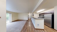 Located in Bothell, WA, this single-floor, 3-bedroom, 2-bathroom unit offers a practical and well-organized layout. The open-concept living and dining area flows seamlessly into the kitchen, which features dark cabinetry, gray stone countertops, stainless steel appliances including a refrigerator and microwave, and durable hardwood-style flooring. The living area extends to a sliding glass door that opens onto a private balcony, bringing in natural light and outdoor views. All three bedrooms are carpeted and finished with warm neutral tones, baseboard heating, and windows with blinds that allow natural light while maintaining privacy. The primary bedroom offers a generous footprint, providing ample space for furnishings. Two full bathrooms mirror each other in design, each featuring a tub-shower combination with white tile surround, dark wood vanity with gray countertop, tile flooring, and chrome fixtures. A hallway connects the bedrooms and bathrooms, with a closet providing additional storage. The unit's consistent use of warm cream walls, rich wood-toned trim, and hardwood-style flooring in common areas creates a cohesive and inviting atmosphere throughout.
