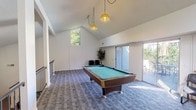This single-floor recreation space is defined by its striking vaulted ceilings that soar to impressive heights, creating an open and airy atmosphere throughout. The room centers around a full-size pool table with a teal felt surface, positioned beneath two vintage-style yellow pendant lights that provide focused illumination and add a retro design touch. Patterned carpet in a neutral blue-grey tone covers the floor, adding texture and comfort underfoot. Natural light filters generously into the space through a large clerestory window positioned high on the wall, as well as through sliding glass doors that lead directly to an outdoor patio. The patio, visible through the expansive glass panels, is furnished with outdoor seating and is surrounded by mature greenery, creating a seamless indoor-outdoor connection. A railed overlook area is visible along one side of the room, offering a view down to an adjoining lounge space below, hinting at the multi-level nature of the broader property. Seating is provided by several black leather chairs arranged around the perimeter of the room, while potted trees add natural warmth to the corners. The clean white walls and open layout make this a versatile and welcoming recreational gathering space.