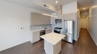 This contemporary single-floor residence showcases a clean, modern aesthetic throughout. The open-concept kitchen is a standout feature, outfitted with light wood grain cabinetry that extends to the ceiling, light grey subway tile backsplash, and a full suite of stainless steel appliances including a gas range, built-in microwave, French door refrigerator, and dishwasher. Light quartz countertops complement the cabinetry, while a central island provides additional workspace and casual seating. A linear pendant light fixture suspended above the island adds a contemporary design element. Dark hardwood-style flooring runs throughout the main living area, creating a warm contrast against the white walls and pale cabinetry. A glass-paneled door leads to a balcony, allowing natural light to fill the space while offering views of the surrounding cityscape. The bathroom features large-format neutral tile flooring, a light wood vanity with an integrated sink, and a walk-in closet area. The overall design palette — neutral tones, clean lines, and modern finishes — creates a cohesive, bright, and functional living environment well-suited for contemporary urban living.