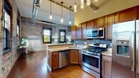 Located in the heart of downtown Buffalo on Main Street, this 1-bedroom, 1-bathroom loft-style residence masterfully blends industrial character with modern finishes. Exposed brick walls are a defining feature throughout, lending warmth and historic charm to every room. Rich hardwood floors run continuously across the space, complementing the building's urban aesthetic.

The open-concept living and dining area is anchored by floor-to-ceiling exposed brick, large multi-pane windows that flood the space with natural light, and soaring ceilings with exposed ductwork that reinforce the loft's industrial identity. The well-appointed kitchen features warm wood cabinetry, granite countertops, a mosaic tile backsplash, stainless steel appliances including a gas range and dishwasher, and stylish pendant lighting hanging from the elevated ceiling. A peninsula with bar seating offers a casual dining option.

A long hallway with exposed brick walls and exposed metal ductwork overhead connects the main living areas to the private bedroom. The bedroom offers high ceilings, a ceiling fan, hardwood floors, and a horizontal window that brings in natural light. The bathroom includes a granite-topped vanity, tile flooring, and access to the hallway. Additional practical spaces include a dedicated laundry room, utility room, and closet, making this a fully functional urban retreat.