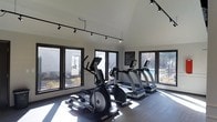 This fitness center offers a clean, modern workout environment designed to keep members motivated and comfortable. The space is bathed in natural light, thanks to expansive floor-to-ceiling windows that line the walls on multiple sides, offering pleasant views of the surrounding outdoor landscape. The bright, airy atmosphere makes the room feel open and inviting throughout the day.

The facility is equipped with a curated selection of cardio machines, including treadmills, an elliptical trainer, and a rowing machine, providing a well-rounded range of cardiovascular workout options. The equipment is arranged thoughtfully to maximize both space and usability, allowing for comfortable movement between stations.

Overhead, a track lighting system runs along the ceiling, providing even, adjustable illumination that complements the natural light streaming in from the windows. The flooring is durable and fitness-appropriate, suited for high-traffic use. The walls are finished in a neutral tone, contributing to the space's clean and focused aesthetic.

An informational posting on the wall suggests the presence of facility rules or guidelines, indicating that this is a shared or amenity-style fitness space. The single-floor layout ensures easy accessibility, and the overall design reflects a practical yet polished approach to fitness facility planning.