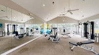 Located in Raleigh, NC, this fitness center offers a spacious, single-floor layout designed to support a full range of workout routines. The room is defined by striking vaulted ceilings that lend an open, airy feel, while multiple ceiling fans ensure consistent airflow throughout. Recessed lighting supplements the generous natural light that pours in through large windows lining the walls, providing bright, welcoming views of the surrounding greenery.

The fitness floor is carpeted and thoughtfully arranged to accommodate both cardio and strength training. On the cardio side, the space features multiple treadmills, elliptical machines, a stationary bike, and a rowing machine, all positioned along the windowed wall for an invigorating outdoor view during workouts. The strength training area includes a full rack of dumbbells, cable machines, a multi-functional weight station, adjustable benches, and a collection of exercise balls for functional training.

A floor-to-ceiling mirror spans one wall, visually expanding the space and allowing users to monitor their form. Entry is provided through elegant French doors with glass panels. The neutral, warm-toned walls and clean white trim create a polished, professional atmosphere. Overall, this fitness center combines practical equipment variety with an inviting architectural design.
