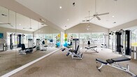 Located in Raleigh, NC, this fitness center offers a spacious, single-floor layout designed to support a full range of workout routines. The room is defined by striking vaulted ceilings that lend an open, airy feel, while multiple ceiling fans ensure consistent airflow throughout. Recessed lighting supplements the generous natural light that pours in through large windows lining the walls, providing bright, welcoming views of the surrounding greenery.

The fitness floor is carpeted and thoughtfully arranged to accommodate both cardio and strength training. On the cardio side, the space features multiple treadmills, elliptical machines, a stationary bike, and a rowing machine, all positioned along the windowed wall for an invigorating outdoor view during workouts. The strength training area includes a full rack of dumbbells, cable machines, a multi-functional weight station, adjustable benches, and a collection of exercise balls for functional training.

A floor-to-ceiling mirror spans one wall, visually expanding the space and allowing users to monitor their form. Entry is provided through elegant French doors with glass panels. The neutral, warm-toned walls and clean white trim create a polished, professional atmosphere. Overall, this fitness center combines practical equipment variety with an inviting architectural design.