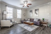 This single-story residence offers a thoughtfully designed open-concept layout that seamlessly connects the living, dining, and kitchen areas. Dark hardwood-style flooring runs throughout the main living spaces, complemented by crown molding and neutral gray tones that lend a cohesive, modern aesthetic. The living room is a comfortable, well-proportioned space anchored by a sectional sofa, a glass-and-wood coffee table, and a ceiling fan overhead.

The kitchen is a standout feature, equipped with white shaker cabinetry, quartz countertops, stainless steel appliances, and a peninsula that provides additional workspace and casual seating. A window above the sink allows natural light to fill the space.

A dedicated office or flex room features dual windows with blinds and sheer curtains, creating a bright and functional work environment. The bedrooms are carpeted and well-lit, each with windows overlooking greenery. The primary bedroom includes a spacious walk-in closet outfitted with wire shelving and hanging rods.

The bathroom features a tub-shower combination with white tile surround and a single-sink vanity. Throughout, the space maintains a clean, contemporary character with practical finishes and a calm neutral palette.