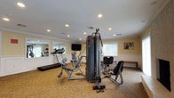 This fitness center is a thoughtfully designed single-floor space that combines functionality with a polished, residential aesthetic. The room features warm yellow walls accented by white wainscoting panels and crown molding, lending the space a refined character uncommon in typical gym environments. Recessed lighting throughout provides consistent, even illumination, complemented by natural light filtering in through multiple windows fitted with vertical blinds.

The main workout area is well-stocked with a range of equipment, including a treadmill, upright stationary bike, elliptical trainer, stair climber, and a multi-station cable weight machine designed to accommodate a variety of strength-training exercises. A wall-mounted flat-screen television is positioned for easy viewing during cardio sessions, and a large mirror along one wall visually expands the space while allowing users to monitor their form.

Durable, patterned carpet covers the floor throughout the space, providing both comfort and traction. A secondary open area adjacent to the main exercise zone offers additional room for stretching or floor exercises. Two white panel doors provide access to connecting areas, and a wall clock keeps users on schedule. Decorative artwork adds a welcoming touch, rounding out an inviting and fully functional fitness environment.