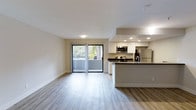 This open-concept studio is thoughtfully designed to maximize its single-floor layout, seamlessly integrating the living and kitchen areas into one cohesive space. The kitchen serves as a functional centerpiece, featuring white cabinetry, dark countertops, stainless steel appliances including a microwave, range, and refrigerator, and a peninsula that provides additional prep space and a natural visual divider within the open floor plan. Recessed lighting illuminates the kitchen area, complementing the abundant natural light that fills the space. Light-toned hardwood-style flooring extends throughout, contributing to a clean, airy aesthetic that enhances the sense of openness. A sliding glass door leads to a private balcony, offering views of lush tropical greenery, including a palm tree, and allowing natural light to pour into the living area. The neutral wall tones and white ceilings further amplify brightness throughout. Vertical blinds on the sliding door provide privacy and light control when needed. Overall, the studio presents a modern, well-appointed environment with efficient use of space.