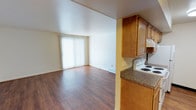This single-floor space features an open-concept layout that connects the kitchen and living area, creating a cohesive and functional environment. The living area is finished with warm hardwood-style flooring that extends throughout, contributing to a clean and inviting atmosphere. A large sliding glass door fitted with vertical blinds allows natural light to fill the space while offering potential access to an outdoor area.

The kitchen is positioned adjacent to the living space and features honey-toned wood cabinetry providing ample storage. Dark granite-look countertops offer a practical work surface, and the kitchen is equipped with a white electric range. An overhead cabinet above the range maximizes vertical storage. The partial wall separating the kitchen from the living area helps define each zone while maintaining the open feel of the overall layout.

The living area is generously proportioned, offering flexible space for furniture arrangement. Neutral white walls and consistent flooring throughout create a bright, unified aesthetic. A floor vent and multiple electrical outlets are conveniently distributed along the walls, adding to the space's practicality.