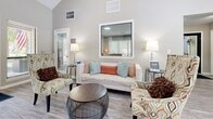 Located at 3844 Battleground Ave in Greensboro, NC, this single-floor space makes a strong impression with its dramatically high ceilings that amplify the sense of openness well beyond its modest footprint. Light gray hardwood-style flooring runs throughout, providing a clean, cohesive foundation for the entire layout.

The living area is warmly furnished with a white sofa, patterned accent chairs, and a round coffee table, arranged around a large interior window that adds depth and visual interest to the space. Soft ambient lighting from table lamps on either side of the seating creates an inviting atmosphere.

A standout feature is the fireplace, which is elegantly framed by a white decorative mantel and marble-style tile surround, with a wall-mounted flat-screen television positioned above. Adjacent to the fireplace, a dining area accommodates a dark wood table and chairs, situated beneath large windows that allow generous natural light to fill the room.

The kitchenette is efficiently designed with white shaker-style cabinetry, granite countertops, a marble tile backsplash, under-cabinet lighting, and a stainless steel compact refrigerator — providing all essential amenities in a streamlined footprint. The overall ambiance is clean, modern, and functional, well-suited for a professional or hospitality-oriented use.