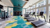 This well-appointed fitness center delivers a comprehensive workout environment designed with both aesthetics and functionality in mind. The space is anchored by a striking geometric-patterned carpet in teal, yellow, and grey tones that energizes the room and defines different workout zones. Bold teal accent walls contrast with crisp white ceilings, creating a dynamic and motivating atmosphere throughout.

The cardio zone features a row of modern treadmills positioned along a wall of large, floor-to-ceiling windows that flood the space with natural light and provide open outdoor views. The strength training area is well-equipped with cable machines, a squat rack, a full dumbbell rack, and a variety of weight benches. Overhead, sleek linear pendant lighting and wall-mounted flat-screen televisions enhance the workout experience.

A dedicated functional training alcove houses a RAX suspension training system, medicine balls, and a punching bag, offering versatility for group or individual training. Mirrored walls expand the sense of space and aid in proper form during exercises. A small utility counter and storage area are also present near the entrance. The soaring ceiling height in select zones adds to the spacious, open feel of the facility.