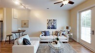 This single-floor residence offers a thoughtfully designed layout with a cohesive, contemporary aesthetic throughout. The kitchen serves as a functional centerpiece, showcasing crisp white shaker-style cabinetry reaching toward the ceiling, dark granite countertops, and a full suite of stainless steel appliances including a range, over-the-range microwave, and refrigerator. A central island provides additional prep space, while light hardwood-style flooring runs the length of the kitchen, adding warmth to the bright, neutral space.

The living area is open and inviting, featuring neutral walls, hardwood-style flooring, ceiling fan with light fixture, and track lighting. The space accommodates generous seating arrangements and is accented by bold artwork, creating a stylish yet comfortable atmosphere.

The bedroom is carpeted and finished in soft, warm tones, with a ceiling fan overhead and a window allowing natural light and outdoor views. A wood-framed dresser with mirror complements the space. A connected bathroom features tile flooring, a tub-shower combination with a decorative curtain, dark granite vanity countertop, and clean, neutral finishes throughout, completing the well-rounded design of this residence.