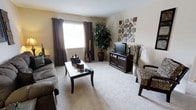 This single-floor space centers around a welcoming living room finished with plush, light-colored wall-to-wall carpeting and warm beige walls that create a neutral, inviting atmosphere. A window dressed with dark chocolate-brown curtains allows natural light to fill the room while providing privacy when needed. The living area is comfortably sized, accommodating a plush sofa, a dark wood coffee table, and a matching entertainment center that houses a flat-screen television. Decorative elements include an ornate metal wall art grouping, potted greenery including a large floor plant, and a zebra-print accent chair that adds a bold visual contrast to the otherwise understated palette. Tall decorative grasses and candle accents contribute additional texture and warmth to the space. The dark espresso-toned furniture pieces provide a rich contrast against the light flooring and walls, lending the room a balanced, layered aesthetic. Overall, the space presents a comfortable and stylishly casual environment suited for relaxed everyday living.