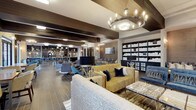 This single-floor communal lounge offers a sophisticated yet welcoming atmosphere defined by its open-concept layout and thoughtful interior design. Exposed dark wood beams run across the ceiling, adding warmth and architectural character to the space, while a prominent multi-bulb chandelier serves as a striking focal point overhead.

The seating area is generously appointed with a curated mix of furniture, including a textured patterned sofa, a bold gold-toned accent loveseat dressed with navy throw pillows, and contemporary swivel chairs in muted tones. A marble-topped coffee table anchors the central gathering area, complementing the eclectic yet cohesive design palette of navy, gold, and grey.

Along one wall, floor-to-ceiling dark shelving units display decorative objects, adding depth and visual interest. A large flat-screen television is positioned centrally, making the space functional for both casual lounging and group viewing.

In the background, a full bar area with pendant lighting and bar-height seating provides a dedicated space for beverages and socializing. The combination of varied textures, layered lighting, and purposeful furnishings creates an inviting, upscale ambiance throughout.