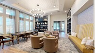 Located in Wesley Chapel, FL, this thoughtfully designed community space at Amberlin Sparrows Edge combines functionality with refined aesthetics across a single, open floor plan. The living area anchors the space with a striking circular chandelier suspended beneath a coffered, shiplap-clad ceiling, casting warm light over a curated seating arrangement of plush boucle swivel chairs, a bold yellow bench nook, and tufted cognac leather wingback chairs positioned beside floor-to-ceiling draped windows that flood the room with natural light and views of the surrounding landscape. The dining area features a substantial wood table with leather upholstered chairs, set atop a patterned area rug over light wood-look flooring that flows seamlessly throughout. Deep navy built-in bookshelves serve as a dramatic focal point, elegantly displaying books, decorative objects, and curated accessories. A secondary built-in shelving unit offers additional display and storage space alongside the cozy bench nook. French doors and large multi-pane windows enhance connectivity to the outdoors, while recessed lighting and warm neutral tones create a welcoming, upscale ambiance suited for both socializing and quiet relaxation.