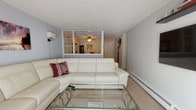Located at 25 English in Manchester, New Hampshire, this single-floor unit offers a well-organized layout with distinct living spaces connected by a functional floor plan. The living room serves as a central gathering area, featuring carpet flooring, a large white sectional sofa, a glass-top coffee table, and a half-wall partition with built-in shelving that opens visually to the dining area beyond. Wall-mounted sconce lighting and decorative artwork contribute to the room's casual, comfortable atmosphere.

The dining room is generously sized, accommodating a full formal dining set with seating for six to eight, complemented by a ceiling fan with a light fixture, a decorative mirror, and a curio cabinet. It connects directly to the galley-style kitchen, which features warm wood cabinetry, tile backsplash, light tile flooring, a full suite of white appliances including a range, dishwasher, and over-range microwave, along with ample counter space.

The unit includes multiple carpeted bedrooms, each with natural window light and baseboard heating. One bedroom features cool gray tones with a modern platform bed, while another offers a warmer aesthetic with rich wood furniture. A third bedroom doubles as a workspace. The bathroom includes a wood-framed vanity with a medicine cabinet and white tile wainscoting.