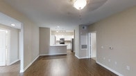 Located at 1935 Northstar Way in San Marcos, this single-story unit offers a thoughtfully designed open-concept layout that seamlessly connects the living area to the kitchen. The living space features rich, dark hardwood-style flooring that extends throughout, complemented by neutral taupe-toned walls and crisp white trim that create a clean, modern aesthetic. A decorative ceiling light fixture anchors the main living area, adding a subtle stylistic touch overhead.

The kitchen is positioned at the rear of the open floor plan and is outfitted with stainless steel appliances, including a range, microwave, and refrigerator. White cabinetry lines the walls and pairs with a light-colored countertop, while a peninsula with bar-height seating provides additional workspace and casual dining space. Track lighting illuminates the kitchen, ensuring a well-lit cooking environment.

Recessed lighting and a visible HVAC vent indicate thoughtful climate control integration throughout the unit. A doorway visible from the main living area leads to additional rooms beyond, suggesting a functional layout that balances open communal space with private accommodations. Overall, the unit presents a contemporary, move-in-ready atmosphere.