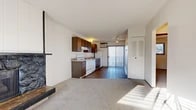 Located at 9310 E Montgomery Ave in Spokane Valley, WA, this single-floor unit offers a practical and comfortable layout suited for everyday living. The open living area greets you upon entry, featuring plush carpeting, a large window that draws in natural light, and a striking stone fireplace framed by vertical wood panel accents — serving as the room's focal point. The kitchen flows seamlessly from the living space, transitioning to durable hardwood-style flooring. It is equipped with dark walnut-toned cabinetry, a white electric range, dishwasher, refrigerator, a double-basin sink, and a wall-mounted AC unit. A sliding glass door leads to an outdoor area, adding both light and ventilation to the kitchen and dining space. The bedroom is generously proportioned with soft carpeting, a ceiling fan, a window overlooking the outdoors, and an expansive set of bi-fold louvered closet doors providing abundant storage. The full bathroom features wood-tone cabinetry with a dark stone-topped vanity, a framed mirror, hardwood-style flooring, and a tub-shower combination. Throughout, the unit maintains a clean, functional aesthetic with a neutral color palette and practical finishes.
