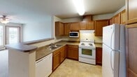 This single-floor space is designed with functionality and comfort in mind, featuring a cohesive neutral color palette accented by warm wood trim throughout. The living area is generously proportioned, finished with plush carpeting underfoot and equipped with a ceiling fan with integrated light fixture. A baseboard heating system runs along the walls, and natural light filters in through windows fitted with blinds, offering views of the surrounding area.

The bedroom is a comfortable, well-lit room also featuring carpeting, a ceiling fan, and a wall-mounted air conditioning unit, providing climate control year-round. Multiple doors lead to closet space and connecting rooms, maintaining a practical flow through the unit.

The kitchen is efficiently laid out in an L-shape configuration, offering ample cabinetry in warm wood tones with dark countertops providing contrast. White appliances include an electric range, built-in microwave, dishwasher, and refrigerator. Tile flooring adds durability to this high-use area.

The bathroom is notably spacious, featuring an accessible roll-in shower with grab bars, a large vanity with wood cabinetry, a wide mirror with overhead lighting, and tile flooring. The accessible design elements make this space both practical and accommodating for a range of needs.
