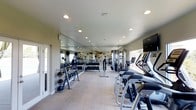 This single-level fitness center is thoughtfully designed to accommodate a full range of workout activities. The space features a clean, open layout with light-toned flooring that contributes to an airy, welcoming atmosphere. Recessed ceiling lights provide consistent, even illumination throughout, while windows along the perimeter allow natural light to filter in, enhancing the overall brightness of the room.

A full-length mirrored wall spans one side of the gym, visually expanding the space and serving a functional purpose for form monitoring during exercise. Motivational text is displayed on the wall behind the mirrors, adding an energetic character to the environment.

The facility is outfitted with a solid selection of equipment, including a row of treadmills and elliptical machines positioned along one side for cardiovascular training. A well-organized free weight section features a tiered dumbbell rack alongside kettlebells and medicine balls for varied strength training. Cable and resistance machines are also present, supporting a comprehensive strength-training experience. A wall-mounted television adds an entertainment option for cardio users. The space is practical, clean, and efficiently arranged.