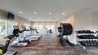 This state-of-the-art fitness facility boasts a spacious layout with multiple exercise areas. The main room features a dedicated cardio section with treadmills and ellipticals, while a separate area houses strength training equipment. Large windows provide abundant natural light, creating an energizing atmosphere. The space is accented with vibrant orange walls, adding a dynamic touch to the otherwise neutral color scheme. High ceilings with recessed lighting enhance the open feel. The center includes specialized equipment like a Solo machine and various weight training stations, catering to a wide range of fitness needs. The textured carpeting throughout adds warmth and absorbs sound, contributing to a comfortable workout environment.