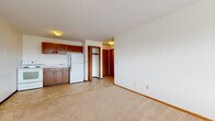 This single-floor unit features an open-concept layout where the living area and kitchen share one continuous space, creating a functional and efficient floor plan. The living area is finished with neutral wall-to-wall carpeting and white walls, providing a clean, versatile backdrop. A large picture window fitted with horizontal blinds allows natural light to fill the room and offers views of the surrounding neighborhood. An under-window HVAC unit provides both heating and cooling.

The kitchen area transitions from carpet to tile flooring and is equipped with warm-toned wood cabinetry above and below the counter, a white electric range, a full-size white refrigerator, and a single-basin sink. The cabinetry provides ample storage for everyday essentials.

A short hallway connects the main living space to the bathroom and a closet. The closet features open hanging space, offering practical storage. The bathroom includes full-height white square wall tile surrounding a soaking tub with an overhead shower, a wall-mounted pedestal sink, a mirrored medicine cabinet, and classic mosaic tile flooring, completing the unit's straightforward and functional design.