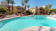 This single-level clubhouse and amenity area showcases a Mediterranean-inspired architectural design, characterized by warm stucco exteriors, tile rooflines, decorative columns, and bold earthy tones that complement the surrounding landscape. At the heart of the outdoor space lies a generously sized, freeform swimming pool with pristine turquoise water, flanked by a spacious pool deck ideal for sunbathing and relaxation. Adjacent to the pool, a dedicated spa features a charming natural rock waterfall feature, adding a tranquil, resort-like ambiance to the environment. Towering palm trees frame the entire area, casting light shade and enhancing the tropical atmosphere under clear, sunny skies. The covered patio area off the clubhouse interior offers a comfortable outdoor lounge with cushioned seating arrangements and pendant lantern lighting, providing a shaded retreat with direct views of the pool through elegant black-framed French doors and windows. Additional outdoor dining areas are furnished with round tables and vibrant red chairs shaded by large umbrellas, perfect for casual gatherings. Well-maintained landscaping with ornamental shrubs and decorative rock borders ties the entire space together, creating a cohesive and inviting community amenity area designed for year-round outdoor enjoyment.
