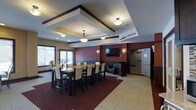 This single-floor space centers around The Goldner Theater, a well-appointed multipurpose room designed for both dining and entertainment. The room features a striking tray ceiling with dark wood trim accents and warm pendant lighting that creates an inviting, polished atmosphere. A large dining table surrounded by high-back upholstered chairs in cream and dark wood provides ample seating for group gatherings. The accent wall, finished in a deep burgundy tone, serves as a focal point and frames a wall-mounted flat-screen television flanked by sconce lighting, lending the space a theater-like ambiance. A cozy seating area with leather chairs is tucked nearby, offering a more relaxed viewing option. Large windows along one wall allow natural light to filter in, brightening the interior. Rich red and neutral patterned carpeting defines the different zones within the room, adding warmth and visual interest underfoot. A doorway at the rear provides access to additional areas of the building. The overall design balances elegance with functionality, making it well-suited for private events, group screenings, or communal dining.