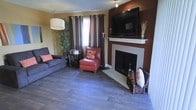 This single-floor space exudes a welcoming and cozy ambiance, anchored by a standout living area that blends comfort with bold design choices. The room is centered around a classic fireplace with a clean white surround and mantel, which serves as a natural focal point and adds warmth to the space. Above the fireplace, a large flat-screen television is mounted against a deep brown accent wall, creating a stylish and functional entertainment area. Rich, dark hardwood flooring runs throughout, grounding the room with a sense of warmth and continuity. The living area is thoughtfully furnished with a plush grey sofa accented with colorful throw pillows, and a vibrant red leather accent chair with a matching ottoman, adding a pop of color that complements the earthy tones of the walls. A small side table and decorative items, including a tall bamboo plant, contribute to the layered and lived-in feel of the space. Natural light filters in through a window dressed with dark brown and tan striped curtains, while a modern floor lamp provides additional ambient lighting. The overall design aesthetic is warm, bold, and inviting, with a thoughtful combination of textures, colors, and furnishings that make this space feel both stylish and comfortable.