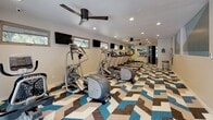 Located at 199 Aegean Way in Vacaville, California, this well-appointed fitness center offers a comprehensive workout environment within a single-floor layout. The space is immediately distinguished by its bold, geometric-patterned carpet in shades of blue, brown, tan, and cream, lending a modern and energetic aesthetic throughout.

The cardio zone is generously equipped with multiple treadmills, elliptical machines, and a stationary bike, arranged in an orderly row along one side of the room to maximize usable floor space. A dedicated strength training area is positioned toward the rear of the facility, featuring a multi-station cable machine and free weights, complemented by a floor-to-ceiling mirror that enhances the sense of openness. Fitness accessories such as stability balls are also available for stretching and functional training.

Overhead, recessed lighting and ceiling fans with sleek dark blades ensure the space remains well-lit and comfortable during workouts. Multiple wall-mounted flat-screen TVs are positioned throughout, offering entertainment while exercising. Horizontal windows along the walls allow natural light to filter in and provide views of the surrounding greenery. Glass double doors at one end of the room open to an outdoor area, further brightening the interior. A custom geometric wall mural adds an artistic focal point, contributing to the facility's clean, contemporary design.